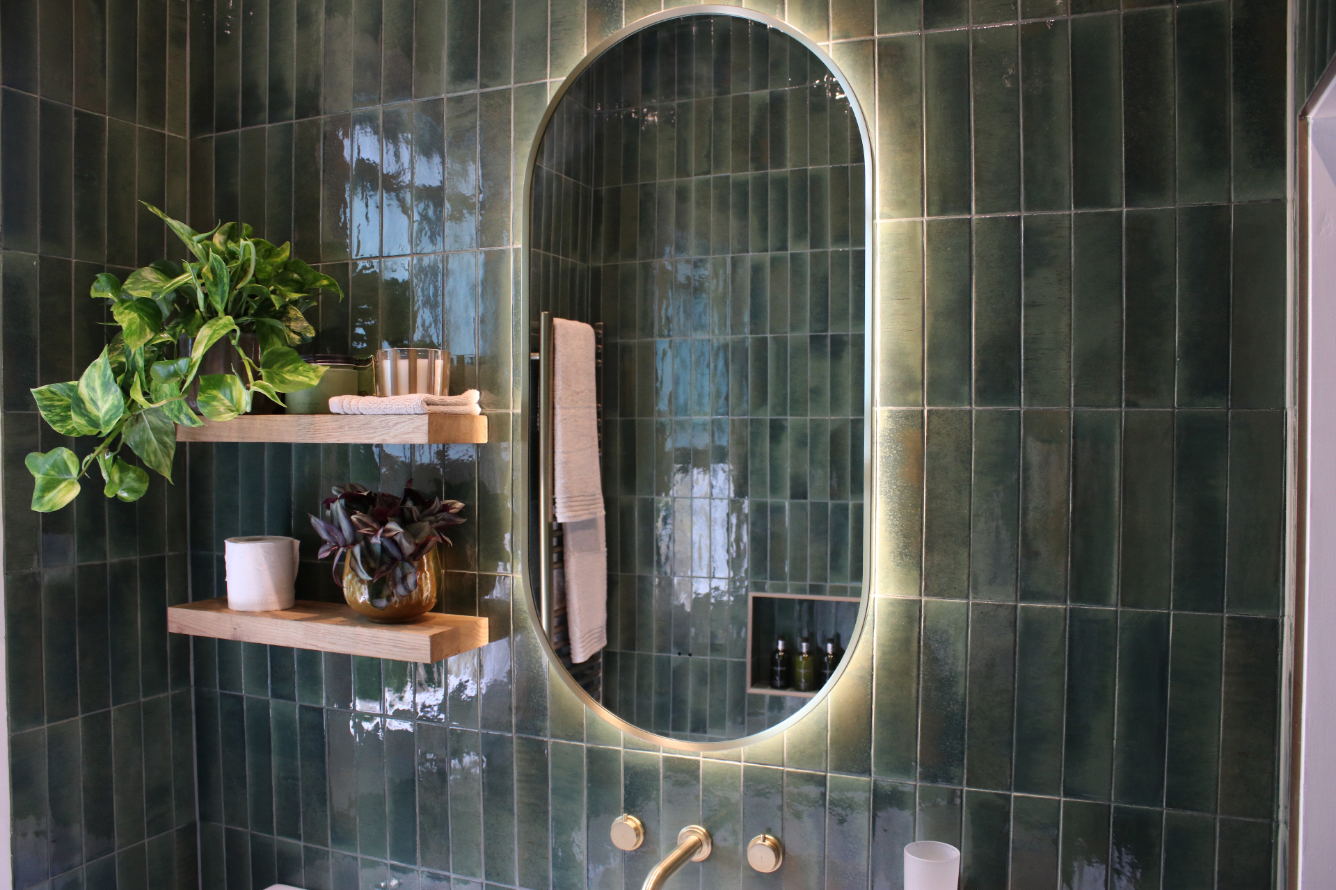 Illuminated arched mirror with brass frame above a brass wall-mounted basin tap, reflected against deep green tiling