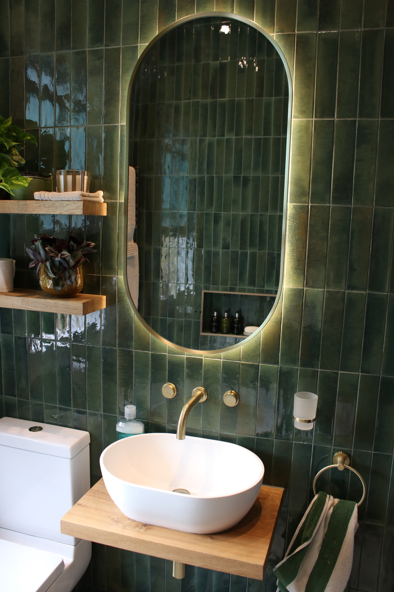 Wall-mounted brass taps and countertop basin beneath the illuminated arched mirror with timber shelving