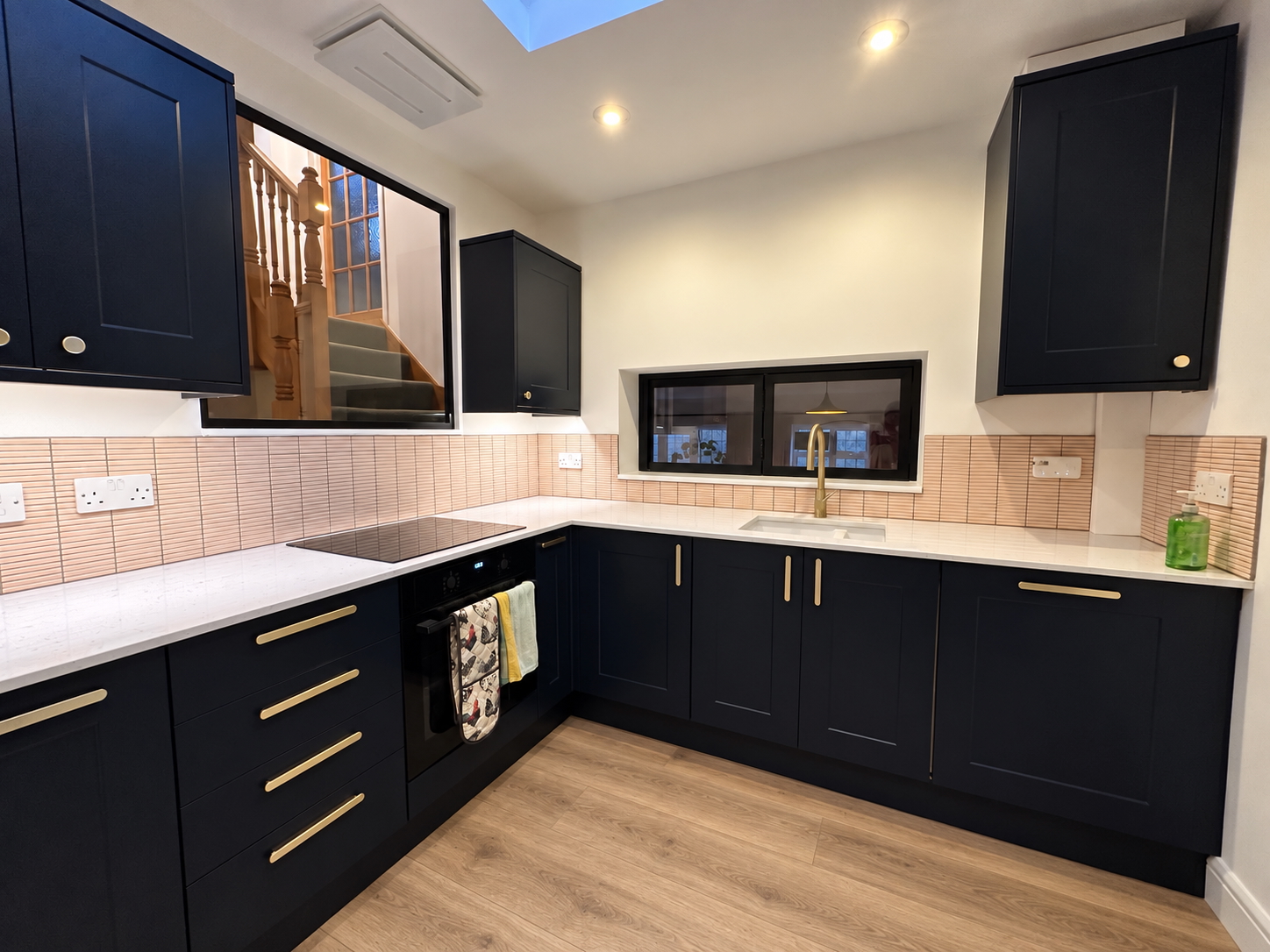 Dark Shaker Kitchen — Searchwood Dark shaker kitchen with pale worktops, brass handles, peach-toned tiled splashback and black-framed internal glazing overlooking the staircase