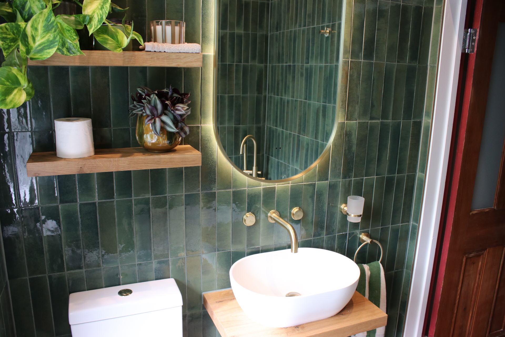 Floating timber vanity with countertop basin, brass taps and oak shelving against deep green tiles
