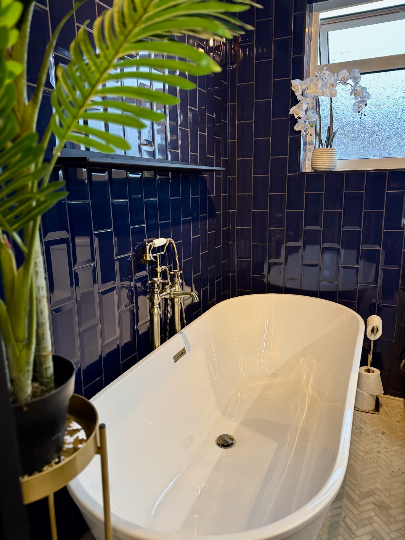 Bath area with black floating shelf, frosted window and brushed brass details above herringbone flooring