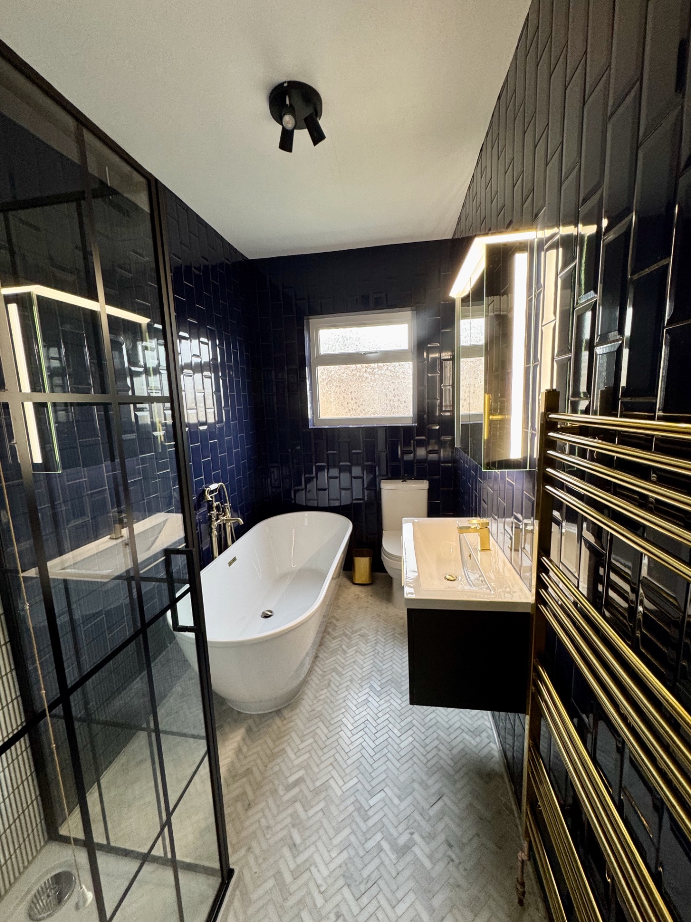 Freestanding bath beneath vertically stacked navy wall tiles with brass fittings and black ceiling light