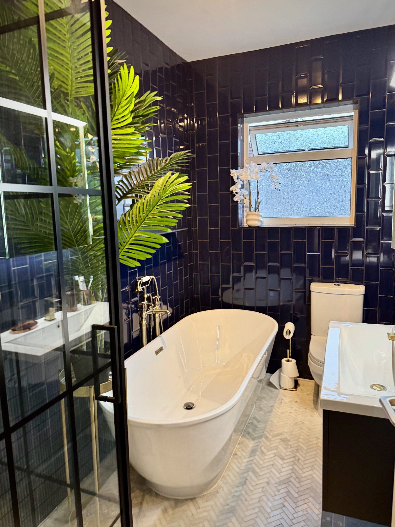 Freestanding bath with floor-mounted brass tap, orchid on the windowsill and navy tiled walls