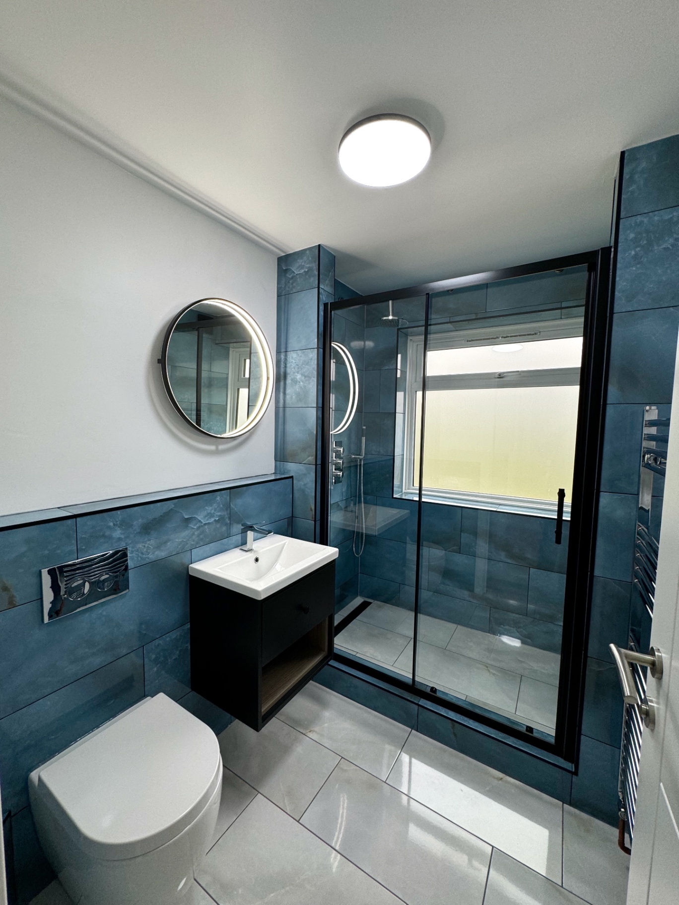 Wall-hung vanity, illuminated circular mirror and modern WC against blue tiled walls